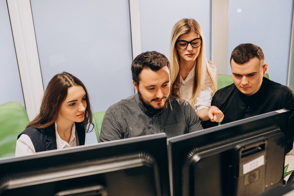 A team of four people collaborating in an office, looking at a computer screen while discussing work.